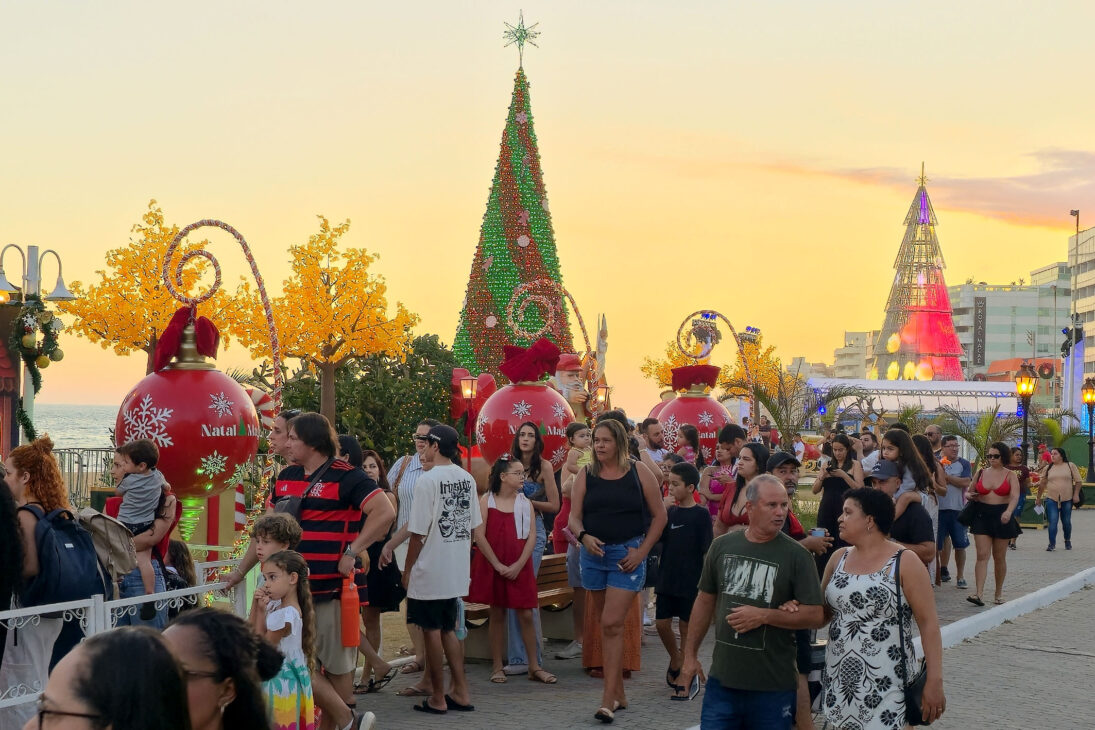 Vila do Noel iluminada na Praia Campista, em Macaé, com famílias caminhando entre árvores de Natal e enfeites gigantes ao pôr do sol