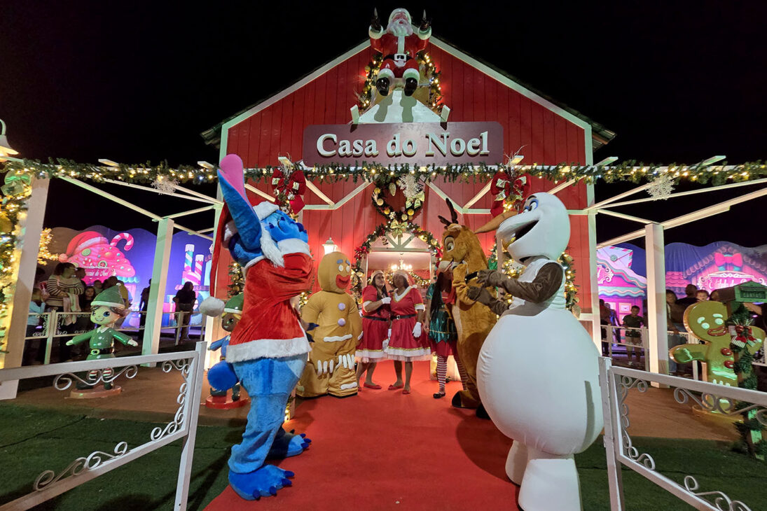 Casa do Noel, com fachada vermelha decorada, Papai Noel inflável no telhado, mascotes no tapete vermelho e luzes de Natal